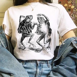 May include: White t-shirt featuring a black and white illustration of two anthropomorphic frogs dancing. The frogs are depicted in a vintage style, with detailed linework and shading. The shirt has the initials "P.B" and the name "JATTAULT" printed on it.