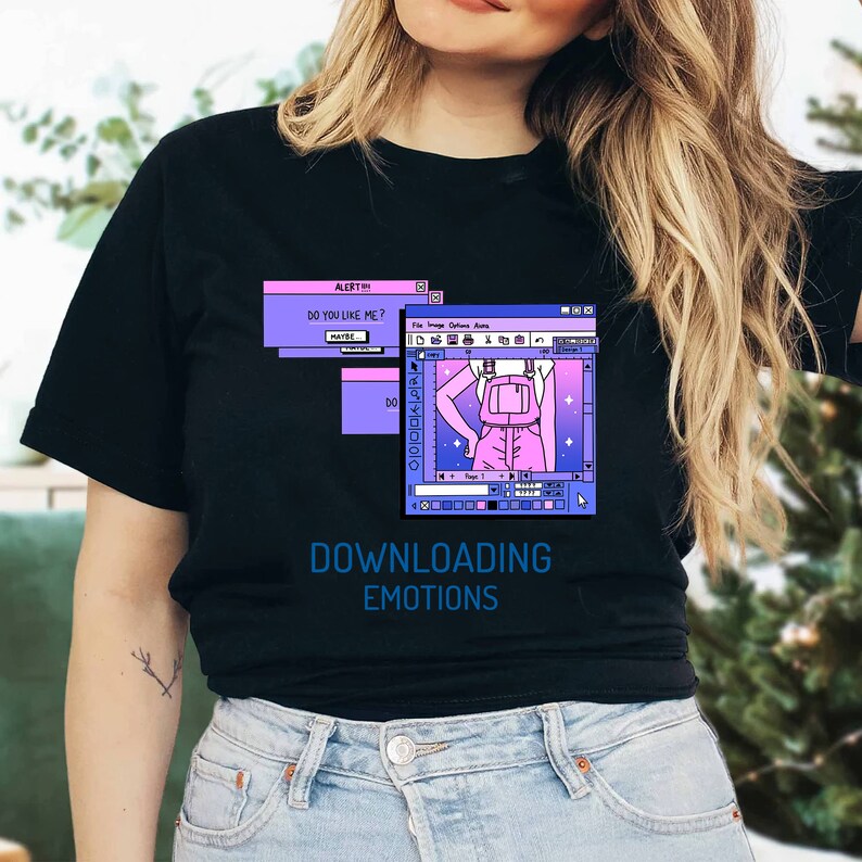 Pu&ograve; includere: T-shirt nero con un design grafico con finestre di computer e la scritta "DOWNLOADING EMOTIONS". Il design include una persona con uno zaino e le parole "DO YOU LIKE ME?" e "MAYBE".
