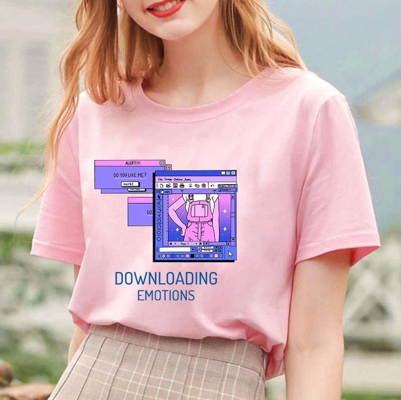 Pu&ograve; includere: T-shirt rosa chiaro con grafica computer retr&ograve;. La grafica include una finestra con una persona con uno zaino e il testo "DOWNLOADING EMOTIONS" e "DO YOU LIKE ME?"