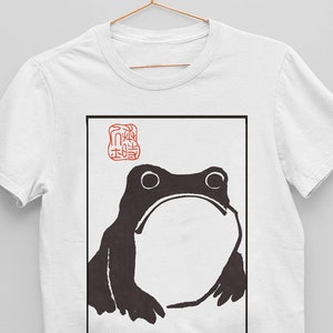 Puede incluir: Camiseta blanca con una ilustración en blanco y negro de una rana. La rana está en un marco cuadrado con un sello rojo que dice "蛙" en japonés.
