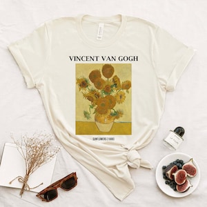 May include: A cream-coloured t-shirt displaying Vincent Van Gogh's "Sunflowers (1888)" painting. The artwork shows a vase of yellow sunflowers. The text "VINCENT VAN GOGH" is above the painting. The t-shirt is folded and knotted.