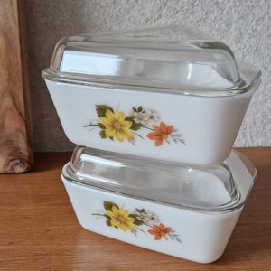 May include: Two white rectangular casserole dishes with clear glass lids. Each dish features a floral design with yellow, orange, and white flowers. The dishes are stacked on top of each other.