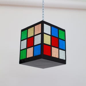 Peut inclure: Un luminaire suspendu en forme de cube noir avec une chaîne. Le cube est composé de 16 petits carrés, chacun d'une couleur différente : vert, or, rose, blanc, bleu et rouge.