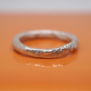 Puede incluir: Un anillo de plata con un diseño texturizado y orgánico. El anillo tiene una superficie rugosa e irregular, lo que le da una apariencia única y artesanal. El anillo está sobre un fondo naranja.
