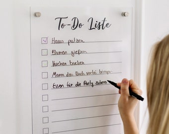 May include: A white acrylic to-do list board with black lines and checkboxes. The text "To-Do Liste" is written at the top of the board. The list includes items such as "Haus putzen", "Blumen gießen", "Kuchen backen", "Mama das Buch vorbei bringen", and "Essen für die Party zubereiten".