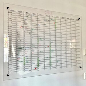 May include: A clear glass dry-erase calendar with a black frame and black push pins. The calendar is divided into months, with each month having 31 days. The calendar is filled with handwritten notes and appointments in black, red, and green ink. The year 2022 is written in pink at the top of the calendar.