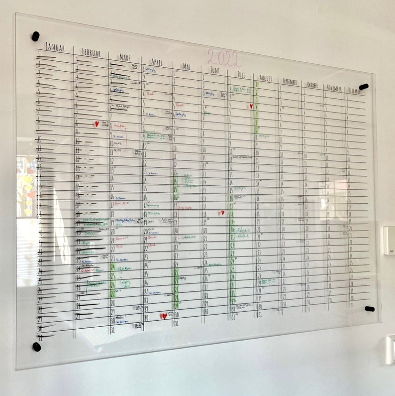 May include: A clear glass dry-erase calendar with a black frame and black push pins. The calendar is divided into months, with each month having 31 days. The calendar is filled with handwritten notes and appointments in black, red, and green ink. The year 2022 is written in pink at the top of the calendar.