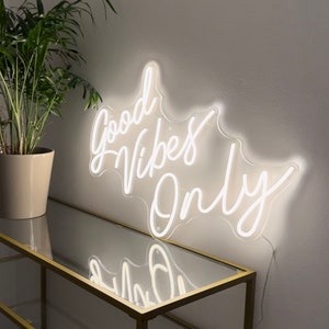 May include: White neon sign with the text "Good Vibes Only" in a cursive font. The sign is mounted on a white wall and is illuminated.