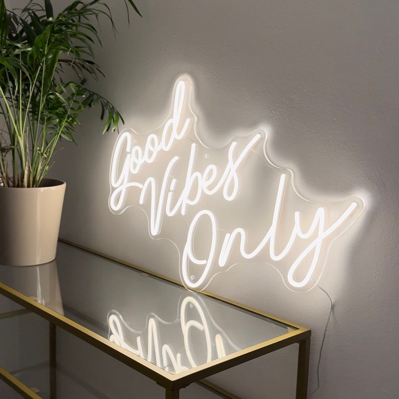May include: White neon sign with the text "Good Vibes Only" in a cursive font. The sign is mounted on a white wall and is illuminated.