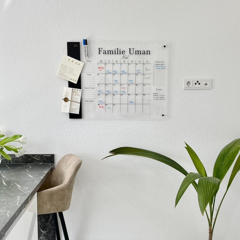 May include: A clear acrylic calendar with the text "Familie Uman Mai" written on it. The calendar is hanging on a white wall with a black magnetic strip and some cards attached to it. There is a potted plant and a chair in the background.