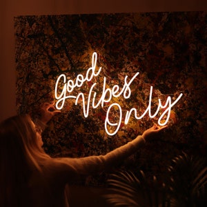 May include: A white neon sign with the text "Good Vibes Only" on a brown and yellow abstract background. A person is reaching up to touch the sign.