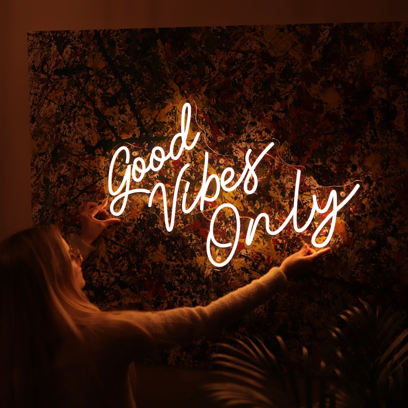 May include: A white neon sign with the text "Good Vibes Only" on a brown and yellow abstract background. A person is reaching up to touch the sign.