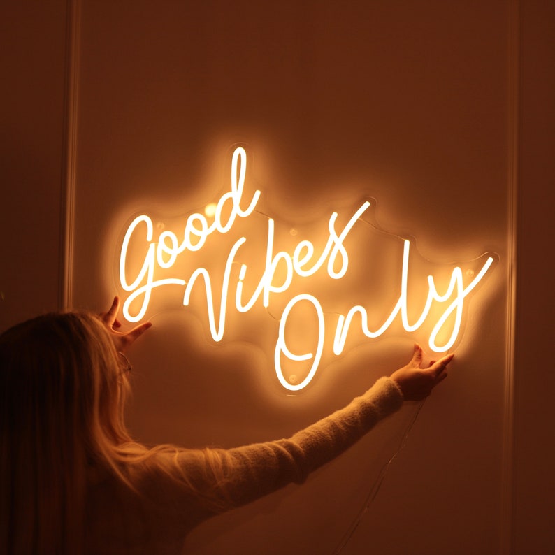 May include: White neon sign with the text "Good Vibes Only" on a white wall. A person is reaching up to touch the sign.