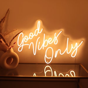 May include: White neon sign with the text "Good Vibes Only" on a beige wall. The sign is lit up and casting a warm glow.