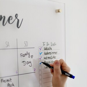 May include: A white dry-erase board with a weekly calendar and a to-do list. The to-do list has checkboxes next to the following items: W&auml;sche, Wohnzimmer, kat zenfu ther, Pass bilder, La Ma. The items are checked off with blue and purple checkmarks. A hand is holding a black marker and checking off the last item on the list.