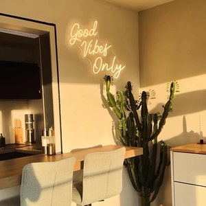 May include: A white neon sign with the text "Good Vibes Only" hanging on a wall with a cactus plant in front of it. The sign is illuminated and casts a warm glow on the wall.