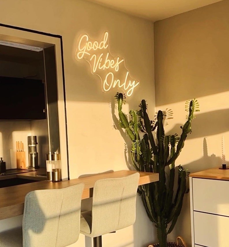 May include: A white neon sign with the text "Good Vibes Only" hanging on a wall with a cactus plant in front of it. The sign is illuminated and casts a warm glow on the wall.