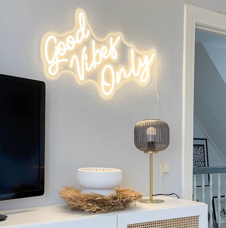May include: White neon sign with the text "Good Vibes Only" on a white wall. A white ceramic candle holder with four candles sits on a table with a brown woven mat. A gold lamp with a dark gray glass shade is on the table.
