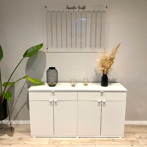 May include: White cabinet with three drawers and three doors. The cabinet has silver handles. A clear acrylic calendar with the text "Familie Sonft" is hanging on the wall above the cabinet. Two small glass candle holders and a black metal lantern are on top of the cabinet.