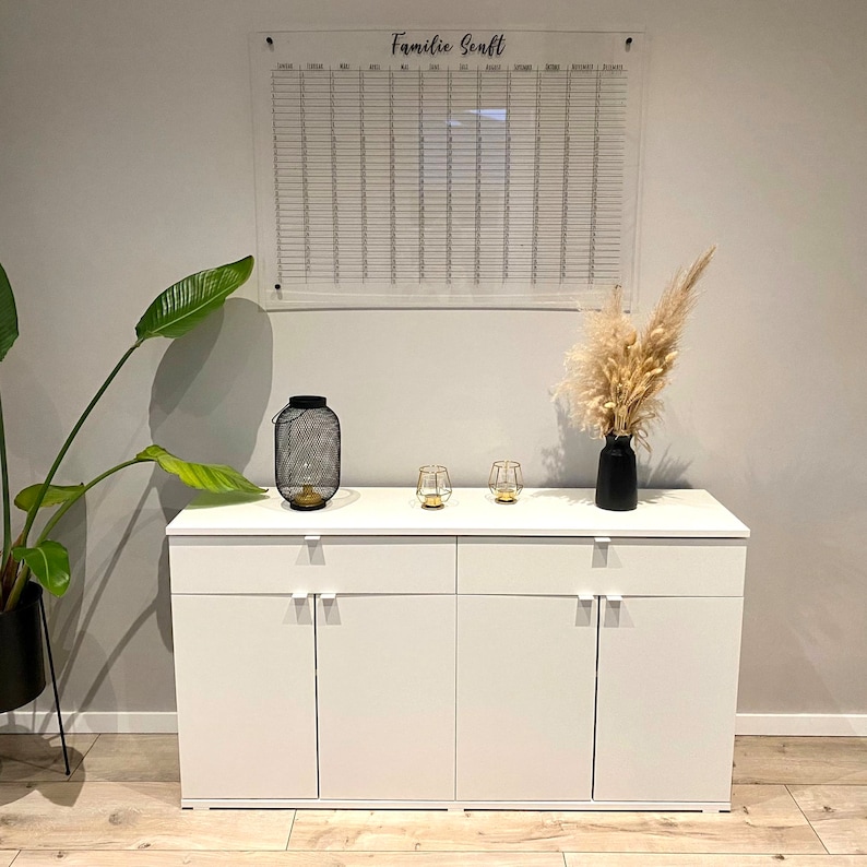 May include: White cabinet with three drawers and three doors. The cabinet has silver handles. A clear acrylic calendar with the text "Familie Sonft" is hanging on the wall above the cabinet. Two small glass candle holders and a black metal lantern are on top of the cabinet.