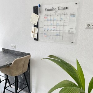 May include: A clear acrylic dry erase calendar with the title "Familie Uman Mai" and a to-do list section. The calendar is mounted on a white wall with a black metal organizer holding cards and pens.