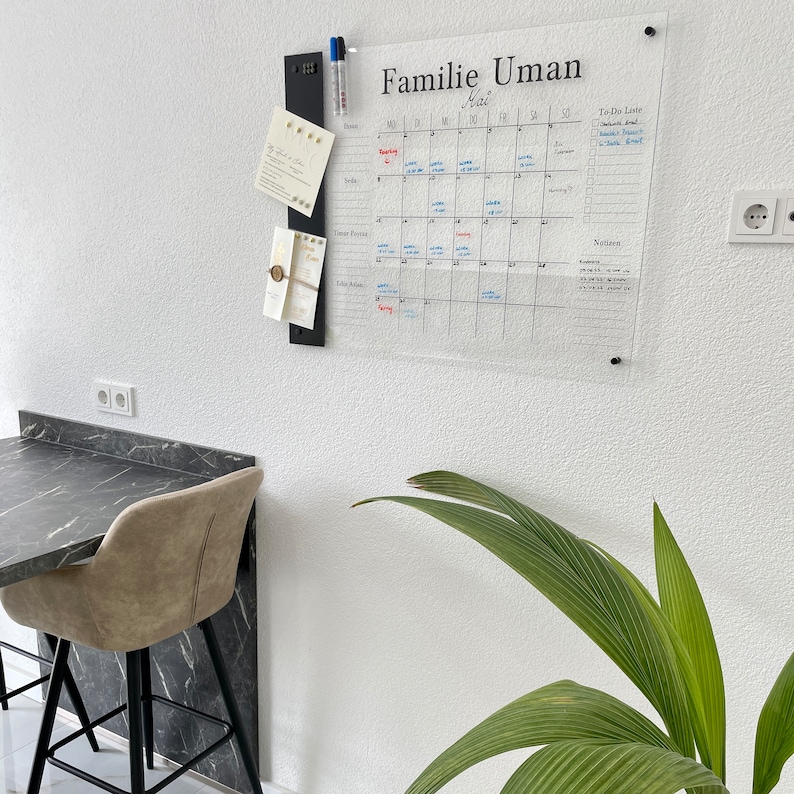 May include: A clear acrylic dry erase calendar with the title "Familie Uman Mai" and a to-do list section. The calendar is mounted on a white wall with a black metal organizer holding cards and pens.
