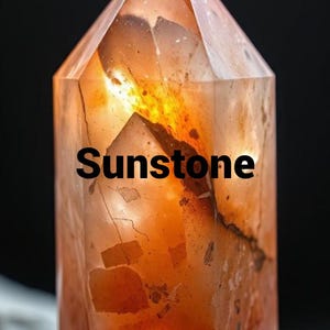 May include: A sunstone crystal with a faceted, pointed top. The translucent stone has warm orange and brown hues, with the word "Sunstone" in black text across the front. The crystal is set against a dark background.