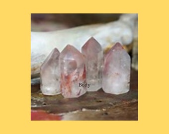 May include: Five clear quartz crystal points with red inclusions. The crystals are arranged in a row on a rough, brown surface. The word "Body" is written on the surface.