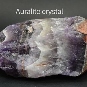 May include: A large, purple and brown Auralite crystal with a rough, natural texture. The crystal has a pointed end and a flat base.