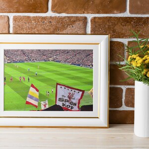 May include: A framed photograph of a soccer game with a green field and a crowd of people in the background. The photo includes a red and white flag with the text "The Original SPION KOP" on it.