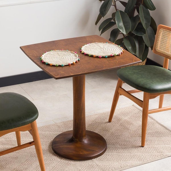 May include: A square wooden pedestal table with two chairs featuring light wood frames, woven cane backrests, and dark green upholstered seats. Two round, cream-coloured crocheted placemats with colourful pom-pom edges sit on the tabletop.