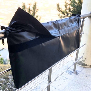 May include: Black fabric cover with a hook and loop fastener, hanging on a metal railing. The cover is designed to protect an object from the elements.