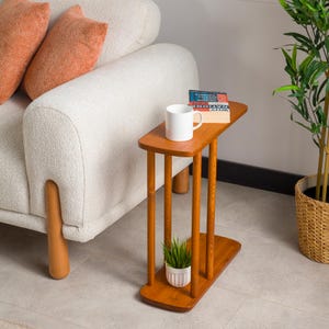 Puede incluir: Una mesa auxiliar de madera con una tapa rectangular y tres patas. La mesa tiene un estante debajo de la tapa. Una taza blanca, una pila de libros y una pequeña planta en maceta están sobre la mesa.