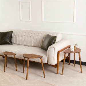 May include: A beige upholstered sofa with vertical channel tufting and two dark olive green throw pillows, accompanied by three small, semi-circular wooden side tables with three thin legs each, arranged in front of the sofa.