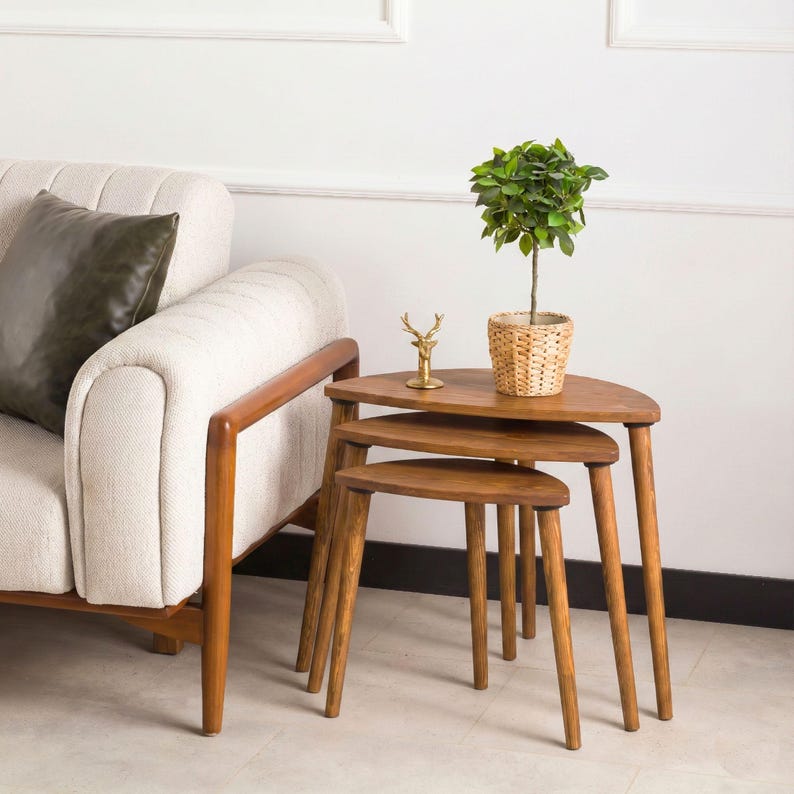 May include: A set of three nesting tables made of medium-toned wood with semi-circular tops and tapered legs. A small potted green plant in a woven basket and a gold-colored deer figurine sit on the largest table surface.
