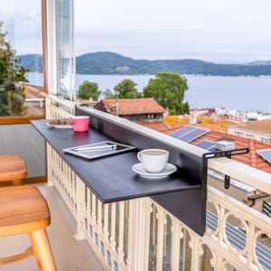 May include: A black metal balcony table with a black tabletop, attached to a white railing. The table has a pink mug, a plate with cookies, a tablet, and a cup of coffee on it. The table is overlooking a scenic view of a body of water and a city.