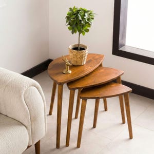 May include: A set of three nesting tables made of medium-toned wood with a smooth finish. Each table features a curved, semi-oval top and three tapered legs. A small potted green plant and a gold-colored deer figurine sit on top.