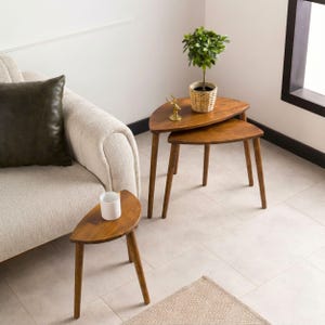 May include: A set of three wooden nesting tables with semi-circular tops and tapered legs. The tables are arranged in a staggered formation, with a small potted green plant and a gold-colored deer figurine placed on the largest table.