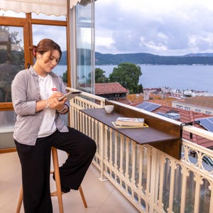 May include: A woman sits on a stool on a balcony, using a tablet and stylus. A wooden table is attached to the balcony railing, with a book and a cup on it. The view from the balcony is of a lake and houses.