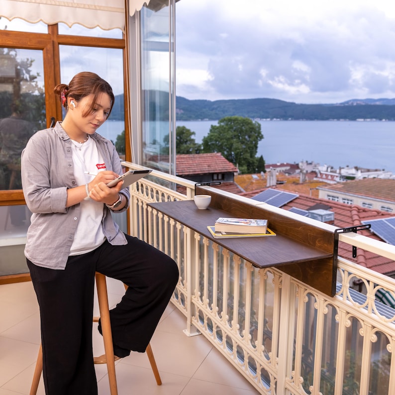 May include: A woman sits on a stool on a balcony, using a tablet and stylus. A wooden table is attached to the balcony railing, with a book and a cup on it. The view from the balcony is of a lake and houses.