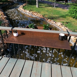May include: A brown wooden shelf attached to a black metal railing with two small potted plants and a black coffee mug on the shelf. The shelf is over a small stream of water.