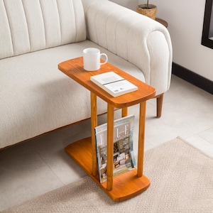 Puede incluir: Una mesa auxiliar de madera con un estante para revistas y un estante. La mesa es marrón y tiene un acabado de madera natural. Está al lado de un sofá blanco y un libro está en el estante superior.