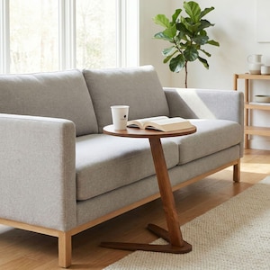 May include: A light grey sofa with a wooden frame and a small, round side table. The table is made of dark wood and has a unique angled leg. A book and a paper cup are on the table.