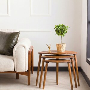 May include: A set of three nested wooden tables with rounded triangular tops and tapered legs. A small potted green plant in a woven basket and a small brass deer figurine are placed on the top surface of the largest table.