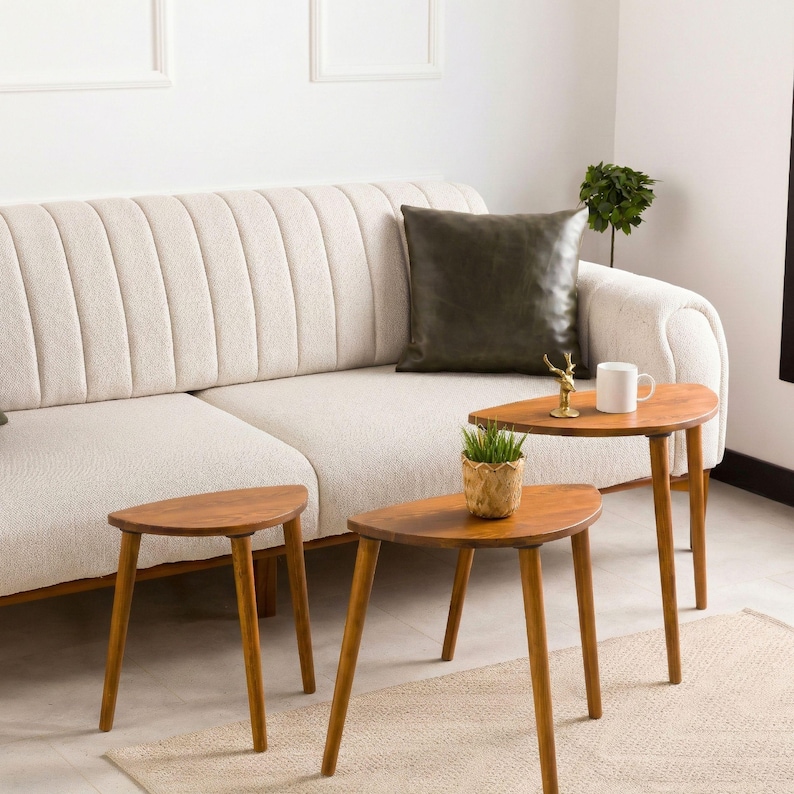 May include: A cream-colored sofa with vertical channel tufting features a dark olive green square pillow. Three semi-circular wooden side tables with tapered legs are arranged in front, holding a small potted plant and a white mug.