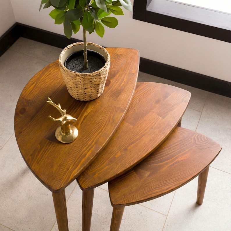 May include: A set of three nesting tables made of medium-brown wood with a smooth finish and curved, triangular tops. A woven basket containing a green plant and a small, gold-colored deer figurine are placed on the largest table.