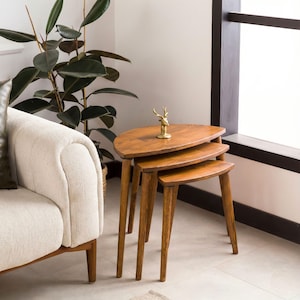 May include: A set of three nesting tables crafted from medium-toned wood, each featuring a smooth, semi-oval tabletop and four tapered legs. A small, gold-colored metal deer figurine stands on the surface of the largest table.