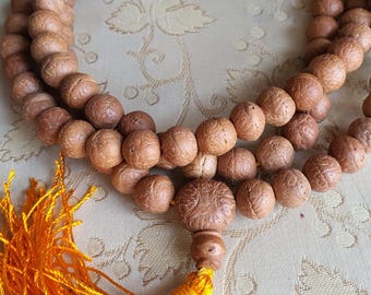 May include: A brown wooden mala bead necklace with a tassel. The beads are round and have a textured surface. The tassel is made of orange thread.