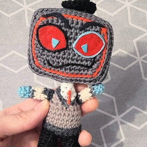Puede incluir: Una muñeca de ganchillo con una cabeza cuadrada de color gris oscuro con borde rojo, grandes ojos rojos con pupilas triangulares azul claro y un sombrero de copa negro con dos antenas de punta roja. El cuerpo lleva un chaleco beige, cuello blanco y pantalón negro.