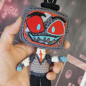 Puede incluir: Una muñeca de ganchillo con una cabeza cuadrada en forma de televisión, un sombrero de copa negro, ojos rojos y una boca azul dentada. El cuerpo lleva un chaleco gris sobre una camisa blanca con corbata roja y pantalones negros.